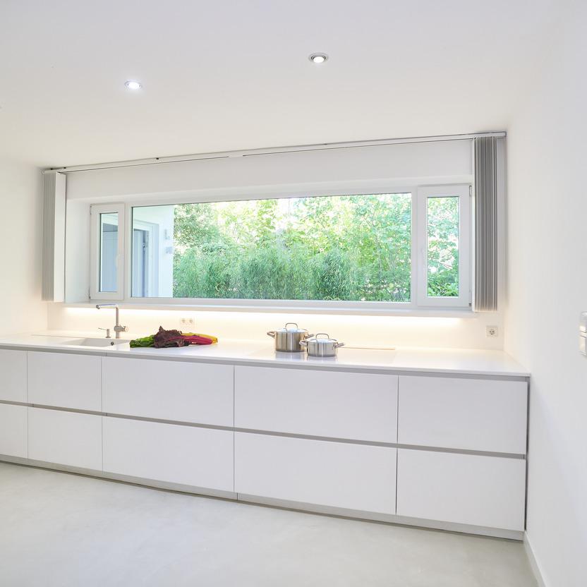 Geradlinige, weiße Küchenzeile nach Maß vor einem großen Fenster mit Blick auf grüne Vegetation.
