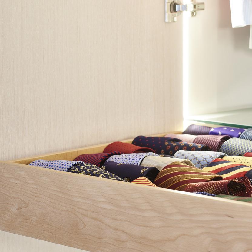 Detailaufnahme einer maßgefertigten Holzschublade mit Fingerzinkung, gefüllt mit ordentlich gerollten Krawatten. Die Schublade ist Teil eines beleuchteten Kleiderschranks mit hellem Innenausbau.