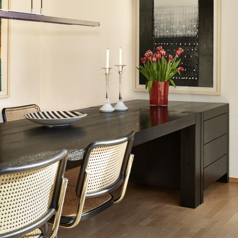 Schwarzer Esstisch nach Maß mit drei seitlich integrierten Schubladen und massiver Holzstruktur. Um den Tisch stehen mehrere Stühle mit Wiener Geflecht. Auf dem Tisch: Tulpenvase, Kerzenleuchter und Keramikschale.