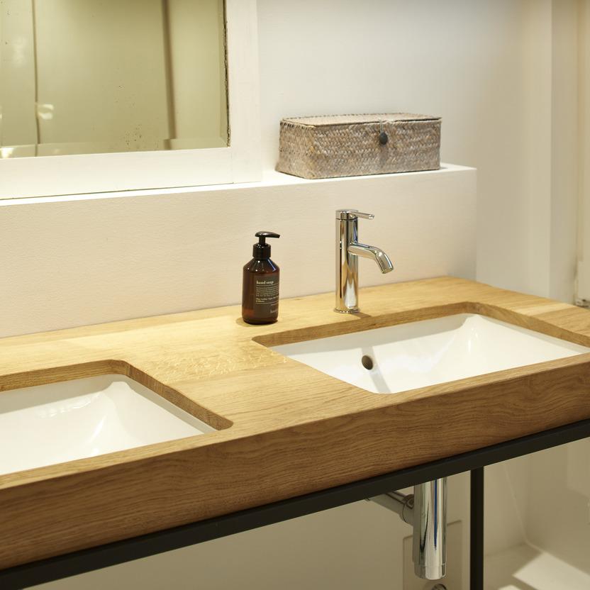 Zwei rechteckige Waschbecken in einer maßgefertigten Holzplatte, begleitet von modernen Chromarmaturen und einem Spiegel mit Holzrahmen, gelegen vor einem Fenster mit Rollo.