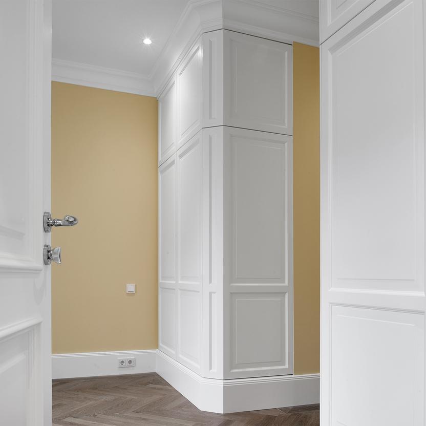 Blick durch eine geöffnete weiße Tür auf einen maßgefertigten Eckschrank mit klassischer Kassettenfront aus unserer Tischlerei. Der Schrank ist exakt in die Raumnische eingebaut, davor gelbe Wandflächen und Fischgrätparkett