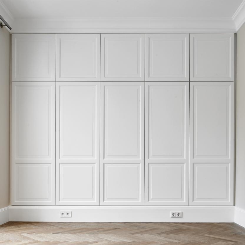 Weißer, maßgefertigter Einbauschrank mit hoher Kassettenfront, bündig in eine Wand integriert. Der Schrank reicht bis zur Decke und steht vor einem hellen Fischgrätparkett, seitlich fällt Licht durch eine große bodentiefe