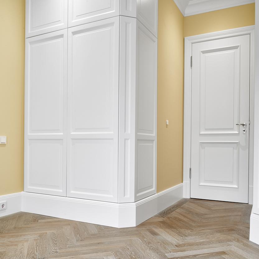 Maßgefertigter, weißer Eckschrank mit klassischer Kassettenfront aus unserer Tischlerei. Der Schrank ist passgenau um die Zimmerecke gebaut und steht vor gelb gestrichenen Wänden mit hellem Fischgrätparkett. Rechts im Bild eine wei&s