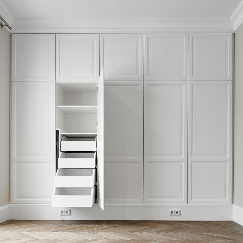 Geöffneter, weißer Einbauschrank nach Maß mit klassischer Kassettenfront. Sichtbar sind mehrere Innenauszüge in unterschiedlichen Höhen sowie ein offenes Regalbrett im oberen Bereich. Der Schrank steht vor hellem Fischgrätp