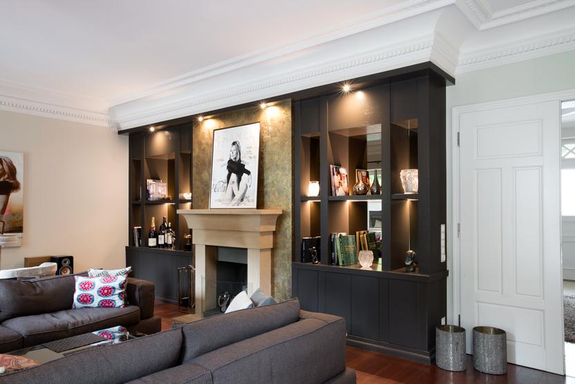 Helles Wohnzimmer mit einer maßgefertigten Wohnwand aus dunklem Holz, eingebaut rund um einen Kamin mit Natursteinverkleidung. Die Regale sind hinterleuchtet und zeigen Bücher, Kunstobjekte und Dekorationen.