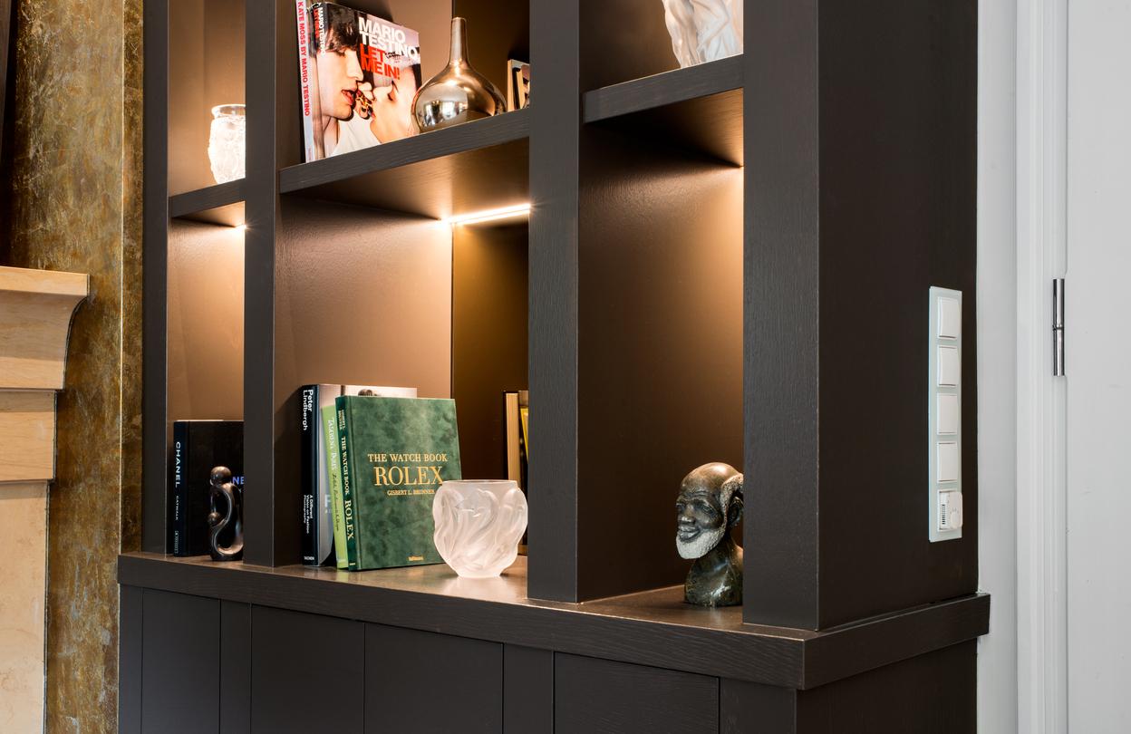 Beleuchtetes Wohnregal nach Maß mit Dekoration und Kunstobjekten Detailansicht eines maßgefertigten, dunklen Wohnregals mit integrierter LED-Beleuchtung. In den Fächern stehen Bücher, Kunstobjekte und Skulpturen. Links grenzt das Regal an einen Kamin mit Natursteinverkleidung, rechts ist ein Lichtschalt